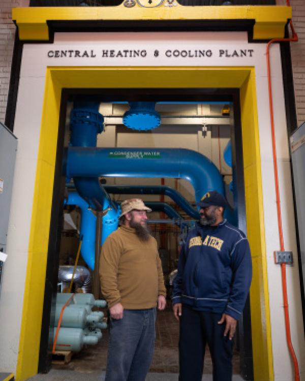 Two men in sturdy work clothes stand in an doorway with the inscription Central Heating and Cooling Plant